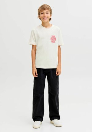 Crème-kleurig katoenen t-shirt met een rode vaasprint op de borst, gecombineerd met losse donkergekleurde broek en witte sneakers.