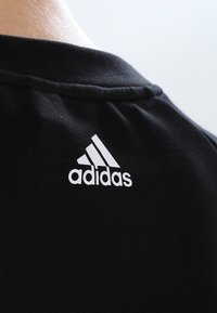 Musta puuvillapaita, jossa on lähikuva takana kaula-aukosta. Paidassa on valkoinen Adidas-logo, jossa on kolme raitaa.