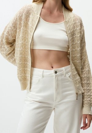 Femme portant un cardigan zippé en dentelle beige sur un crop top blanc et un pantalon blanc taille haute, posant avec une main dans le dos.
