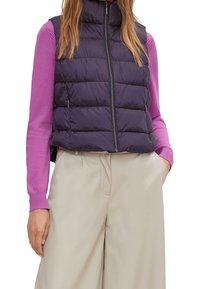 Marineblauer Steppweste mit gestepptem Design, hohem Kragen und Reißverschluss, über einem pinkfarbenen Langarmoberteil und beigefarbenen weiten Hosen. 