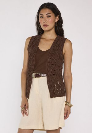 Frau trägt eine braune ärmellose Häkelweste über einem braunen Tanktop mit beigen Shorts, goldene Creolen, Armbänder und Ringe.