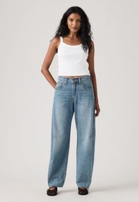 Γυναίκα με μακριά μαύρα μαλλιά φοράει λευκό αμάνικο crop top, ανοιχτό μπλε ψηλόμεσο παντελόνι με φαρδιά μπατζάκια και μαύρα πλεκτά παπούτσια, στέκεται μπροστά σε απλό φόντο.