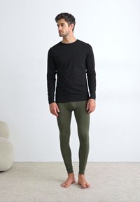 Long-sleeve black shirt paired with fitted olive green leggings. Soft, stretchable fabric with a streamlined design. Model barefoot, standing.