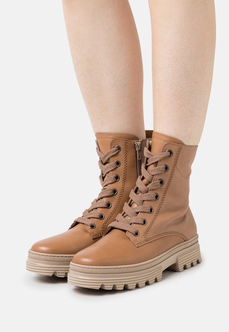 Bottes cavalières en cuir tanné avec lacets, semelles épaisses beiges, portées sur des jambes nues sur fond blanc.