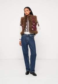 Hnedá kožúšok s kvetinovým vzorom, dlhá biela vyšívaná košela, modré vysoké džínsy a čierne topánky. Jednoduché pozadie.