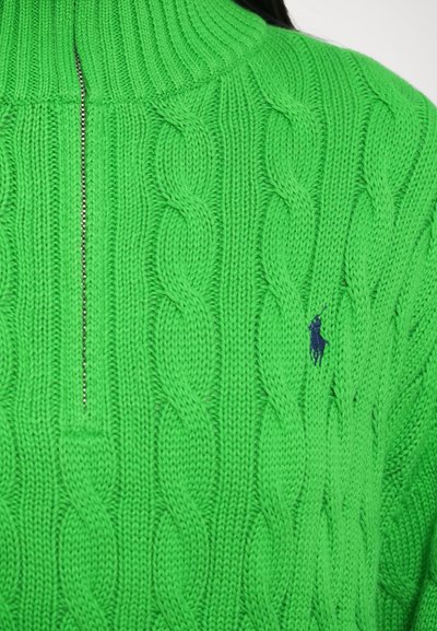 Pletený sveter v jasne zelenej farbe s vrkočovým vzorom, vysokým golierom a štvrťovým zipsom; na hrudi je malý námornícky modrý logotyp.
