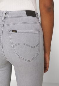 Vaqueros de mezclilla gris con un diseño ajustado, que cuentan con una etiqueta de cuero negro en la cintura y un patrón de costura curvado en los bolsillos traseros.