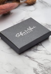 Scatola di gioielli nera con texture, con un logo in rilievo argentato che legge "Sofia Milani Jewels", posata su una superficie di marmo.