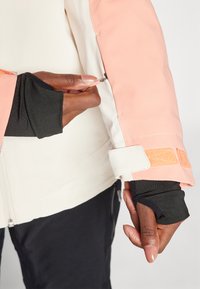 Giacca leggera con un design pesca e bianco, dotata di polsini regolabili e tasca con zip sulla manica, evidenziando diverse texture.