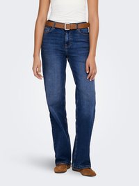 Mėlyni džinsai su platėjančiomis apačiomis, vidutinio aukščio juosmuo, rudas odinis diržas ir šviesios siūlės. Modelis dėvi rudus zomšinius batus.