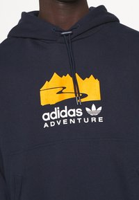 Námořnická modrá mikina s kapucí se siluetou oranžové hory a řeky. Text "adidas ADVENTURE" v bílé barvě pod designem. Smíšená textilní textura.