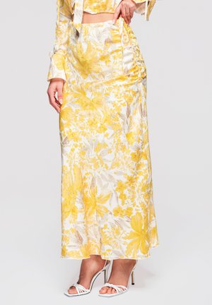 Femme portant une jupe longue maxi jaune et blanche à fleurs avec un détail froncé, associée à des sandales blanches à talons hauts et à lanières, debout devant un fond uni.