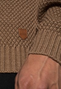 Maglione lavorato a maglia in marrone chiaro. Tessuto spesso con polsini e orlo a coste. Presenta una piccola etichetta in pelle marrone con testo in rilievo.