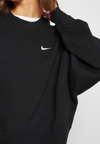 Kvinna som bär en svart Nike-tröja med vitt logo, med ena handen bakom nacken, vit skjortkrage synlig under.