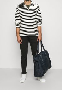 Maglietta a maniche lunghe a righe bianca e nera con colletto a zip, abbinata a pantaloni neri e scarpe da ginnastica bianche. Tiene una grande borsa a tracolla blu navy.