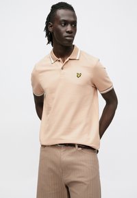 Világos barackszínű, texturált finisű pamut pólóing sárga pillangólógóval a mellkason és kontrasztos csíkos gallérral.