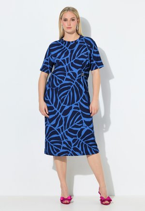 Frau trägt ein midi Kleid mit blau-schwarzem Blättermuster, kurzen Ärmeln und pinkfarbenen Sandalen mit hohen Absätzen und Schleifen, steht vor einer weißen Wand.