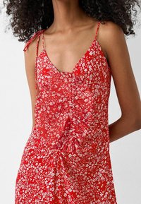 Czerwona sukienka w kwiaty z białymi wzorami, na ramiączkach spaghetti, z dekoltem w kształcie litery V oraz marszczeniem na środku i u dołu. Wykonana z lekkiego materiału.