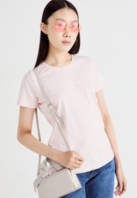 T-shirt de algodão rosa claro com um logótipo branco discreto, combinado com uma bolsa cinza com um detalhe em nó e jeans de ganga azul.