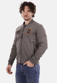 Giacca bomber grigia con zip frontale, due tasche anteriori, toppe ricamate sul petto e polsini e orlo a coste.