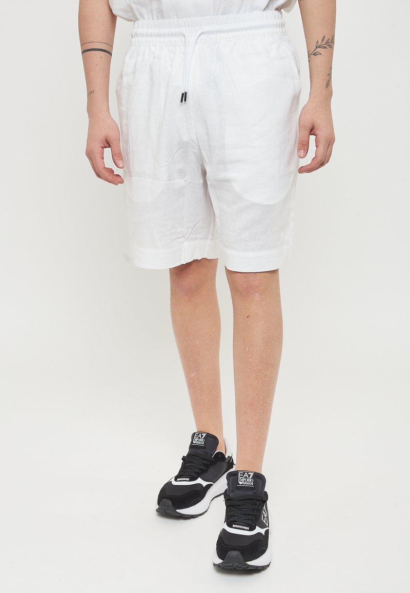 Pantaloni corti bianchi in lino con una cintura elastica e cordino. Indossati con sneaker nere con accenti bianchi e branding. Tessuto liscio.