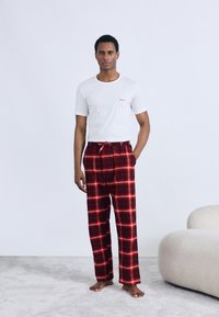 Bijela majica kratkih rukava s crvenim logotipom "HUGO", uparena s crvenim i crnim kariranim pidžama hlačama. Model stoji bos na svijetlom tepihu.