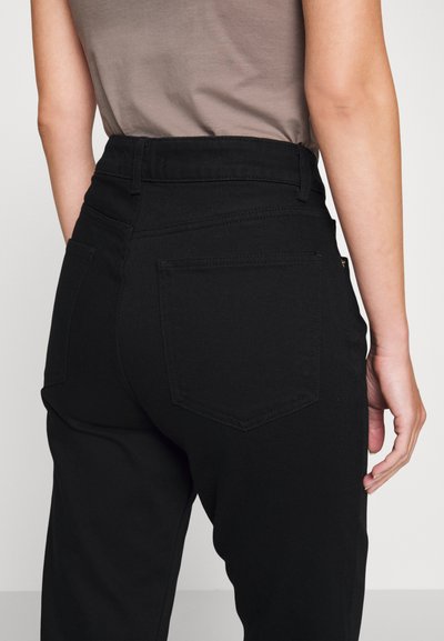 Pantalones de mezclilla negros de cintura alta, con dos bolsillos traseros, costuras visibles y una textura suave. El brazo está posicionado cerca de la cintura.
