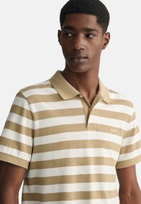 Camisa polo de rayas en beige y blanco con cuello, mangas cortas y pequeño logo de la marca en el pecho. Material de algodón con acabado texturizado.