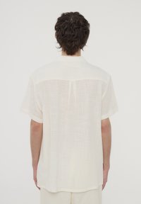 Camicia a maniche corte di colore bianco sporco, realizzata in tessuto leggero con un motivo testurizzato, dotata di un colletto semplice e una vestibilità rilassata.