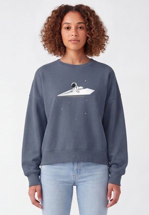 Frau mit lockigem Haar, die einen grauen Sweatshirt mit einer Illustration eines Astronauten auf einem Papierflieger trägt, der im Weltraum schwebt.