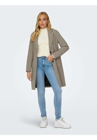 Cappotto lungo a scacchi in beige e marrone, abbinato a un dolcevita bianco, jeans skinny azzurri chiari e sneakers bianche.