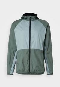 Veste légère à capuche avec un design bicolore, composé d'un haut vert foncé et d'une partie inférieure vert clair. Elle possède une fermeture éclair frontale et des poignets élastiques.