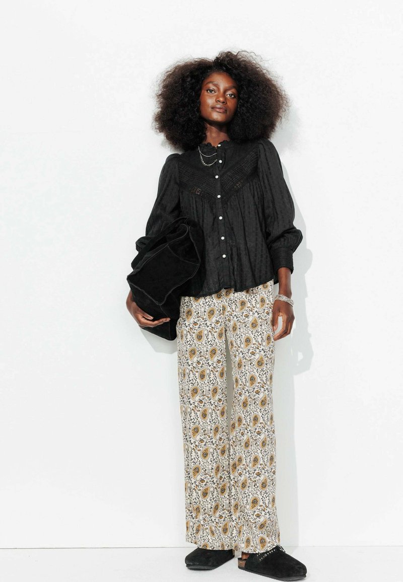 Blouse noire avec des manches bouffantes et patte de boutonnage, associée à un pantalon ample à motif cachemire en blanc, jaune et noir. Chaussures à enfiler noires.