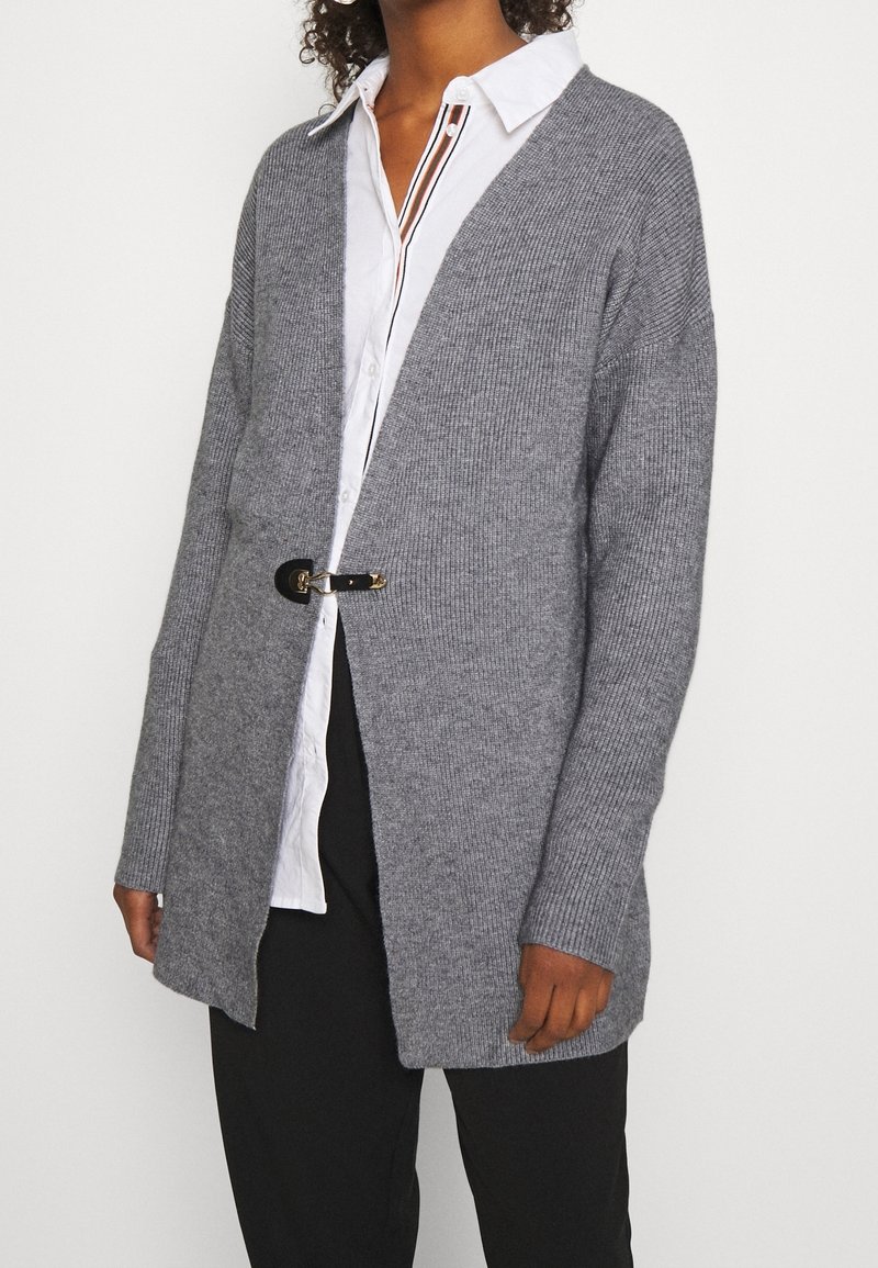 Cardigan gris côtelé avec une ouverture frontale, doté d'une fermeture à boucle dorée. Porté sur une chemise blanche et un pantalon noir.