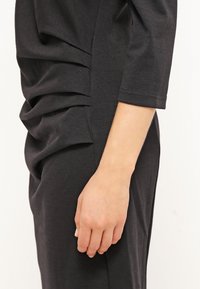 Vue de profil d'une personne portant une robe sombre froncée à manches trois-quarts, montrant le bras et la main détendue sur un fond clair.