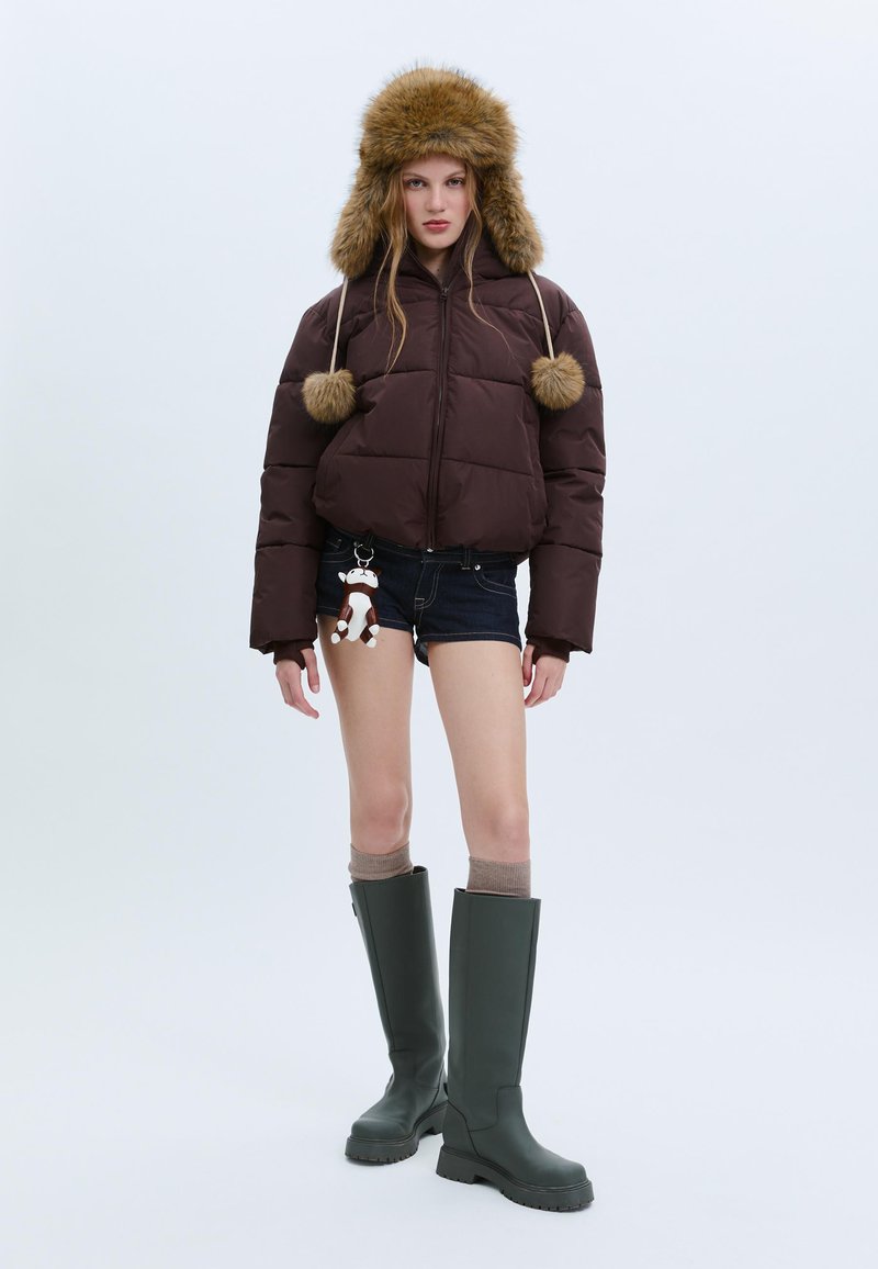 Hnědá péřová bunda s chlupatou kapucí, která má třásně. Nositelná s denimovými šortkami a zelenými gumovými holínkami po kolena. Neutrální pozadí.