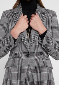 Femme portant un blazer à carreaux noirs et blancs avec une jupe assortie, ajustant les revers du blazer, avec un col roulé noir et un collier en or.