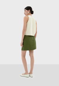 Gehaakte crèmekleurige mouwloze top in combinatie met een groene rok, met een rits aan de achterkant en lichtgekleurde instappers. Over het geheel gladde texturen.