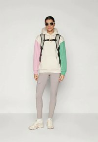 Lietus jaka krēmkrāsā ar rozā un zaļām piedurknēm, apvienota ar gaiši pelēkiem legingiem un baltām kedām, kā arī melnu mugursomu.