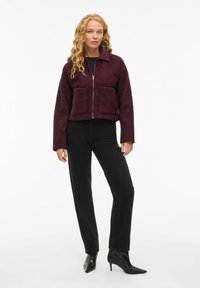 Veste cropped bourgogne en tissu texturé, avec deux poches avant et une fermeture éclair, assortie à un pantalon noir et des escarpins noirs à bout pointu.