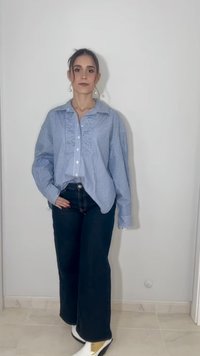 Νεαρή γυναίκα στέκεται, φοράει μια φαρδιά μπλε ριγέ πουκαμίσα, σκούρο τζιν με φαρδιά πόδια, λευκά μποτάκια και σκουλαρίκια με πέρλες, μπροστά από έναν απλό φωτεινό τοίχο.
