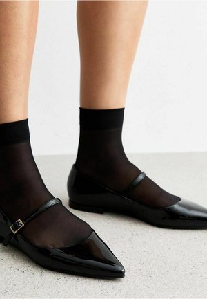 Zwarte lakleren ballerina's met puntige neuzen en een smalle enkelband, gecombineerd met doorschijnende zwarte sokken, op een lichtgrijze ondergrond.