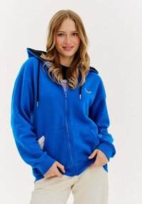 Niebieska bluza z kapturem na zamek błyskawiczny, z czarnym wnętrzem, z długimi rękawami, przednimi kieszeniami i małą grafiką rzęs na klatce piersiowej.