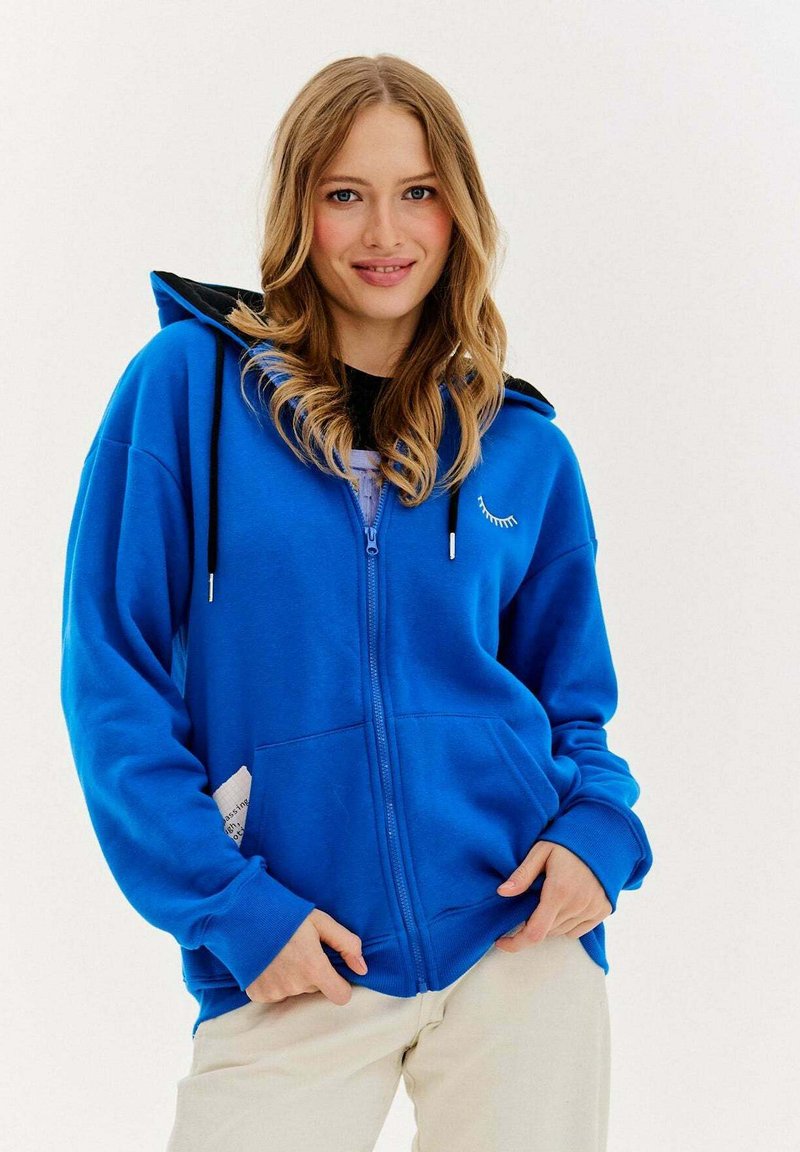 Niebieska bluza z kapturem na zamek błyskawiczny, z czarnym wnętrzem, z długimi rękawami, przednimi kieszeniami i małą grafiką rzęs na klatce piersiowej.