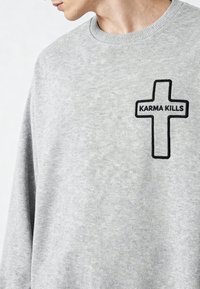 Personne portant un sweatshirt gris clair avec une croix entourée de noir et le texte "KARMA KILLS" sur la partie supérieure gauche de la poitrine.