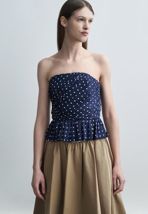 Kvinne med rett brunt hår iført en skulderløs marineblå topp med polkaprikker og et beige skjørt foran en ensfarget grå bakgrunn.