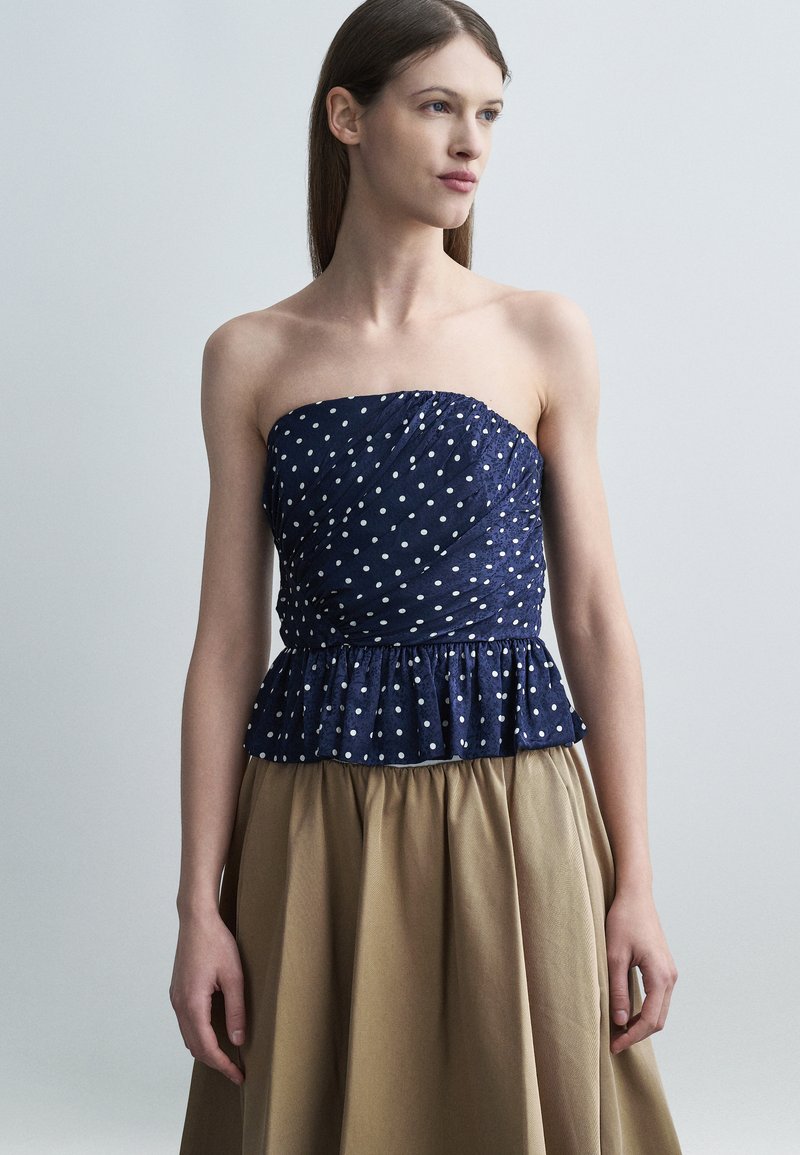 Γυναίκα με ίσια καστανά μαλλιά που φοράει μπλε ναυτικό τοπ με πουά χωρίς τιράντες και μπεζ φούστα σε απλό γκρι φόντο.