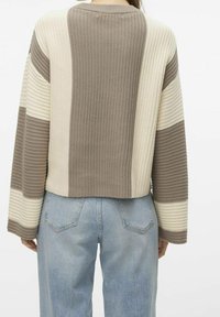 Pull en tricot avec des rayures beige, crème et grises. Présente une texture côtelée et une coupe décontractée et courte. Porté avec un jean bleu clair.