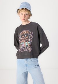 Gray cropped sweatshirt with colorful "Ripcurl" graphic, paired with light blue denim jeans. The model wears a light blue bucket hat.