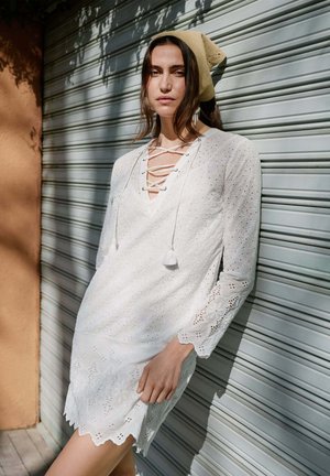 Nainen seisoo metalliverhoilun edessä valkoisessa silkkipainetussa mekossa, jossa on nyörityskaulus ja beige huivi päässä, auringonvalo heittää varjoja seinälle.